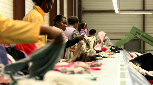 L’image montre plusieurs personnes travaillant dans un centre de tri textile. Elles sont alignées le long d’un tapis roulant, sélectionnant et triant des vêtements d’occasion. L’environnement industriel, l’éclairage fluorescent et la disposition du tri indiquent un processus structuré de recyclage ou de revalorisation des textiles.