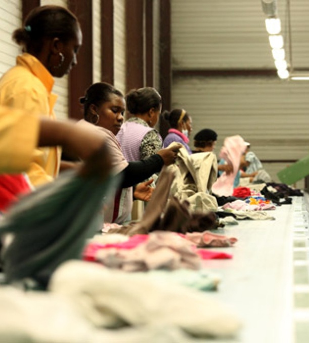 L’image montre plusieurs personnes travaillant dans un centre de tri textile. Elles sont alignées le long d’un tapis roulant, sélectionnant et triant des vêtements d’occasion. L’environnement industriel, l’éclairage fluorescent et la disposition du tri indiquent un processus structuré de recyclage ou de revalorisation des textiles.