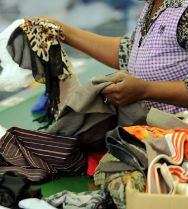 Une personne trie des vêtements d’occasion sur un tapis roulant. Elle porte un tablier violet à motifs vichy sur une chemise à imprimé léopard. Elle manipule divers textiles colorés, dont des tissus à motifs et des tissus unis. L’environnement évoque un centre de tri de vêtements recyclés.