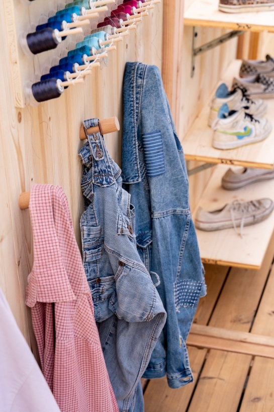 Une scène chaleureuse et artisanale avec des vêtements suspendus sur des crochets en bois, dont une salopette et une veste en denim patchée. En arrière-plan, des étagères en bois exposent des baskets usées, tandis qu’un assortiment de bobines de fil de différentes couleurs ajoute une touche créative à l’ensemble.