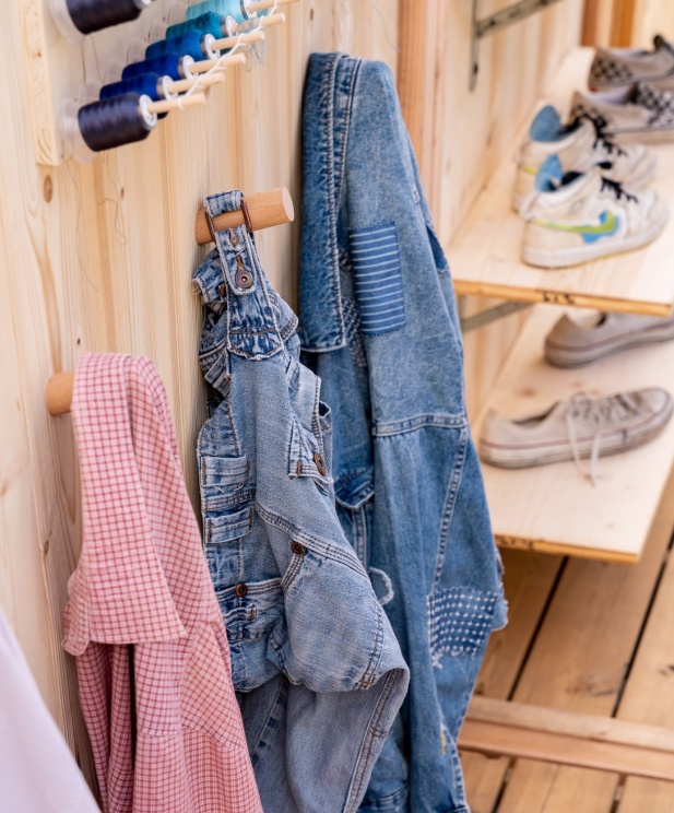 Une scène chaleureuse et artisanale avec des vêtements suspendus sur des crochets en bois, dont une salopette et une veste en denim patchée. En arrière-plan, des étagères en bois exposent des baskets usées, tandis qu’un assortiment de bobines de fil de différentes couleurs ajoute une touche créative à l’ensemble.