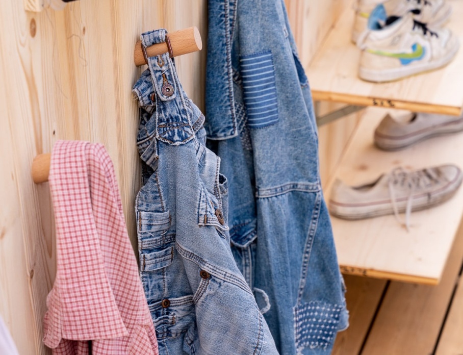 Une scène chaleureuse et artisanale avec des vêtements suspendus sur des crochets en bois, dont une salopette et une veste en denim patchée. En arrière-plan, des étagères en bois exposent des baskets usées, tandis qu’un assortiment de bobines de fil de différentes couleurs ajoute une touche créative à l’ensemble.