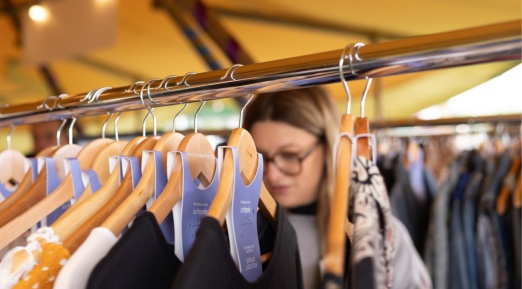 Friperie sous un chapiteau : des vêtements suspendus sur des cintres en bois, une main fouille parmi les pièces tandis qu'une jeune femme aux lunettes explore le portant.