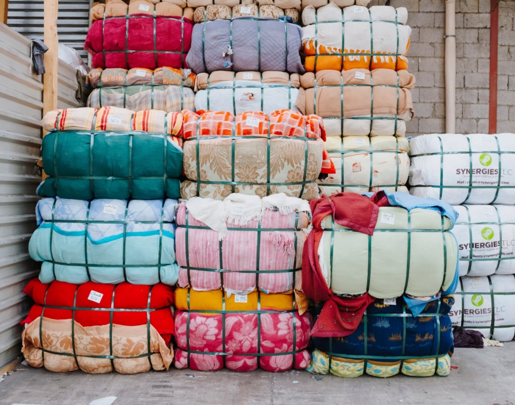 Empilement de ballots de textiles colorés compressés et maintenus par des sangles vertes. Les tissus présentent divers motifs et couleurs. Certains ballots portent des étiquettes, et à droite, des paquets blancs marqués "SYNERGIES LLC" sont visibles. L’environnement est un entrepôt avec un mur en parpaings et une structure métallique.
