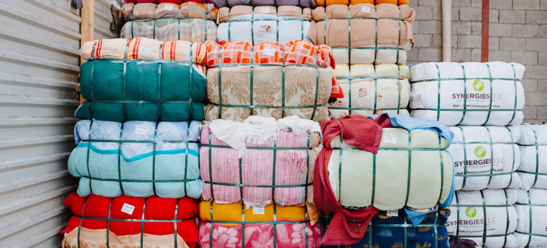 Empilement de ballots de textiles colorés compressés et maintenus par des sangles vertes. Les tissus présentent divers motifs et couleurs. Certains ballots portent des étiquettes, et à droite, des paquets blancs marqués "SYNERGIES LLC" sont visibles. L’environnement est un entrepôt avec un mur en parpaings et une structure métallique.