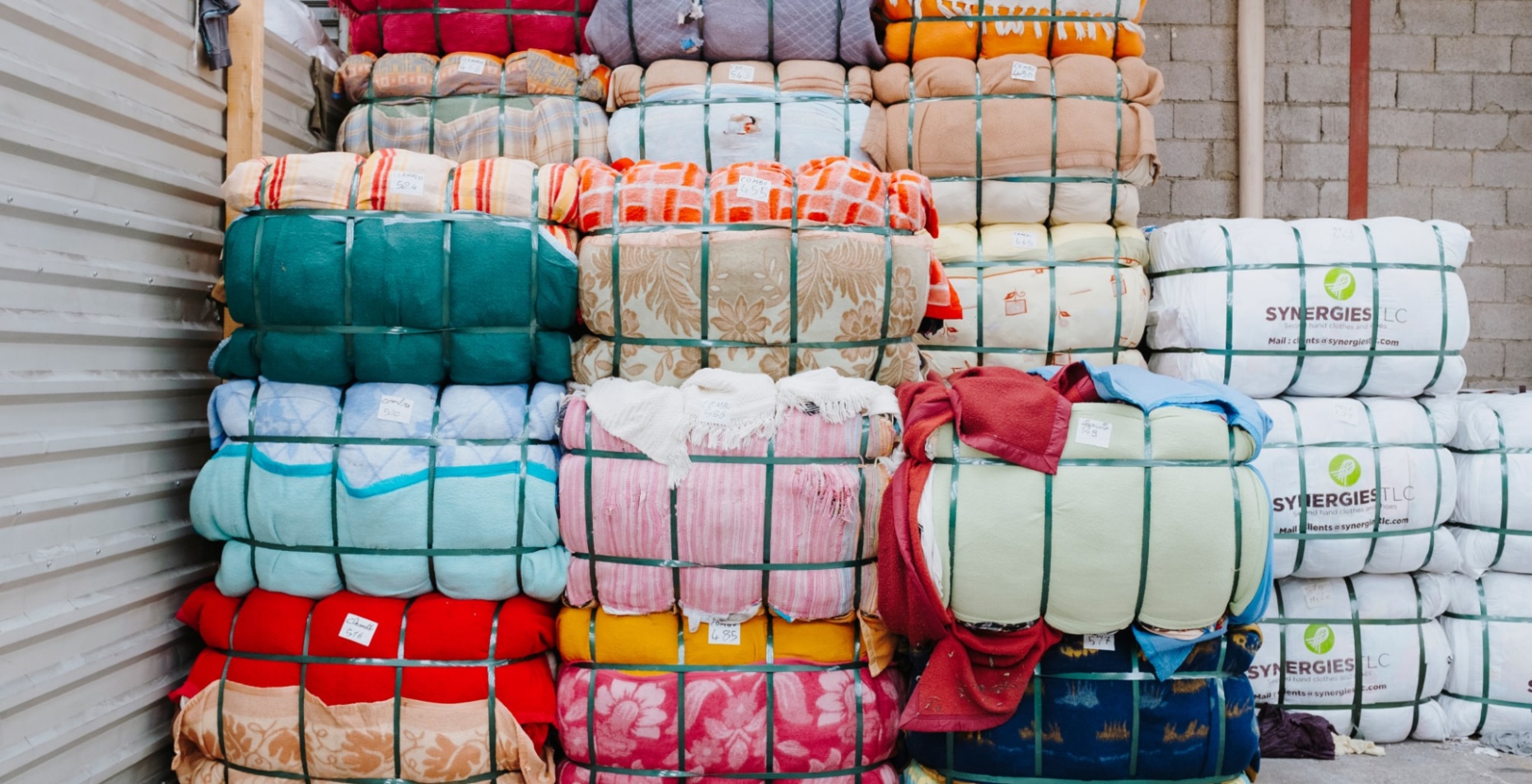 Empilement de ballots de textiles colorés compressés et maintenus par des sangles vertes. Les tissus présentent divers motifs et couleurs. Certains ballots portent des étiquettes, et à droite, des paquets blancs marqués "SYNERGIES LLC" sont visibles. L’environnement est un entrepôt avec un mur en parpaings et une structure métallique.