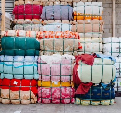 Empilement de ballots de textiles colorés compressés et maintenus par des sangles vertes. Les tissus présentent divers motifs et couleurs. Certains ballots portent des étiquettes, et à droite, des paquets blancs marqués "SYNERGIES LLC" sont visibles. L’environnement est un entrepôt avec un mur en parpaings et une structure métallique.