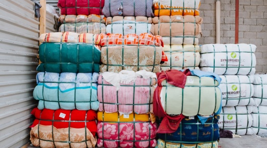 Empilement de ballots de textiles colorés compressés et maintenus par des sangles vertes. Les tissus présentent divers motifs et couleurs. Certains ballots portent des étiquettes, et à droite, des paquets blancs marqués "SYNERGIES LLC" sont visibles. L’environnement est un entrepôt avec un mur en parpaings et une structure métallique.