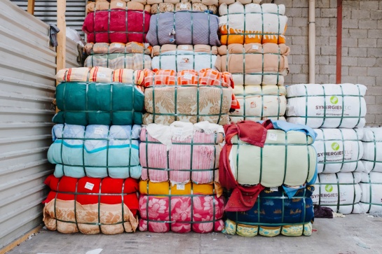 Empilement de ballots de textiles colorés compressés et maintenus par des sangles vertes. Les tissus présentent divers motifs et couleurs. Certains ballots portent des étiquettes, et à droite, des paquets blancs marqués "SYNERGIES LLC" sont visibles. L’environnement est un entrepôt avec un mur en parpaings et une structure métallique.