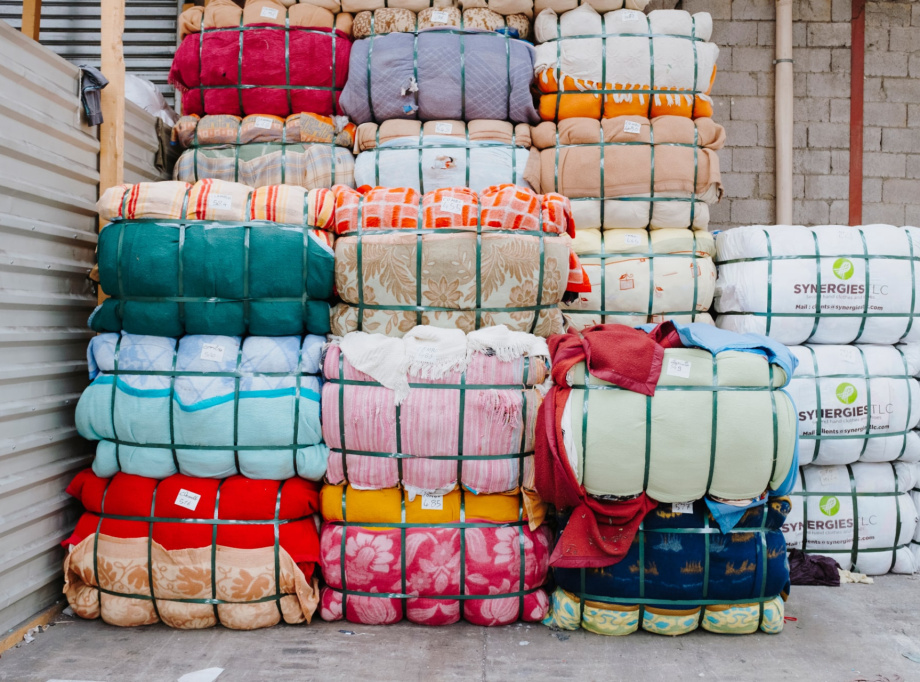Empilement de ballots de textiles colorés compressés et maintenus par des sangles vertes. Les tissus présentent divers motifs et couleurs. Certains ballots portent des étiquettes, et à droite, des paquets blancs marqués "SYNERGIES LLC" sont visibles. L’environnement est un entrepôt avec un mur en parpaings et une structure métallique.