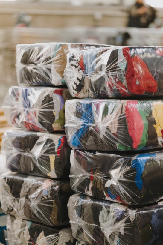 Gros plan sur des ballots de vêtements compressés et emballés dans du plastique transparent. Les textiles sont de différentes couleurs et textures. En arrière-plan, un environnement industriel flou avec une personne visible.