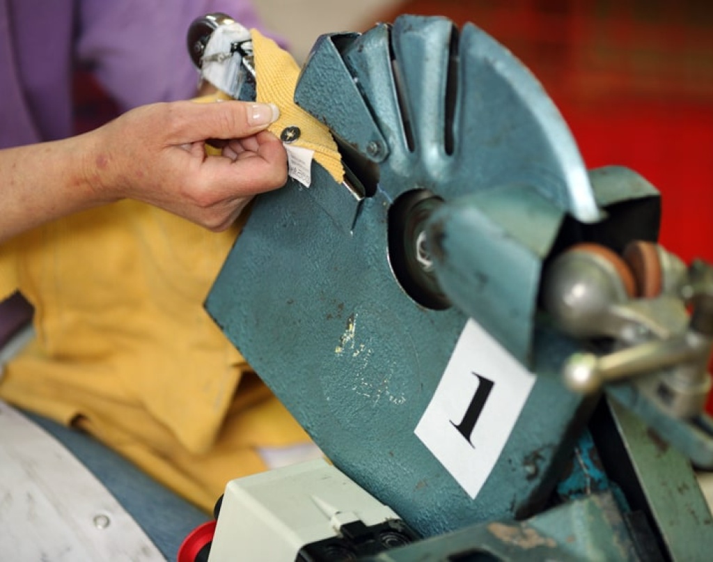 Une personne utilise une machine industrielle en métal bleu avec un mécanisme rotatif pour retirer un bouton pression d'un tissu jaune. La machine est étiquetée avec le chiffre "1".