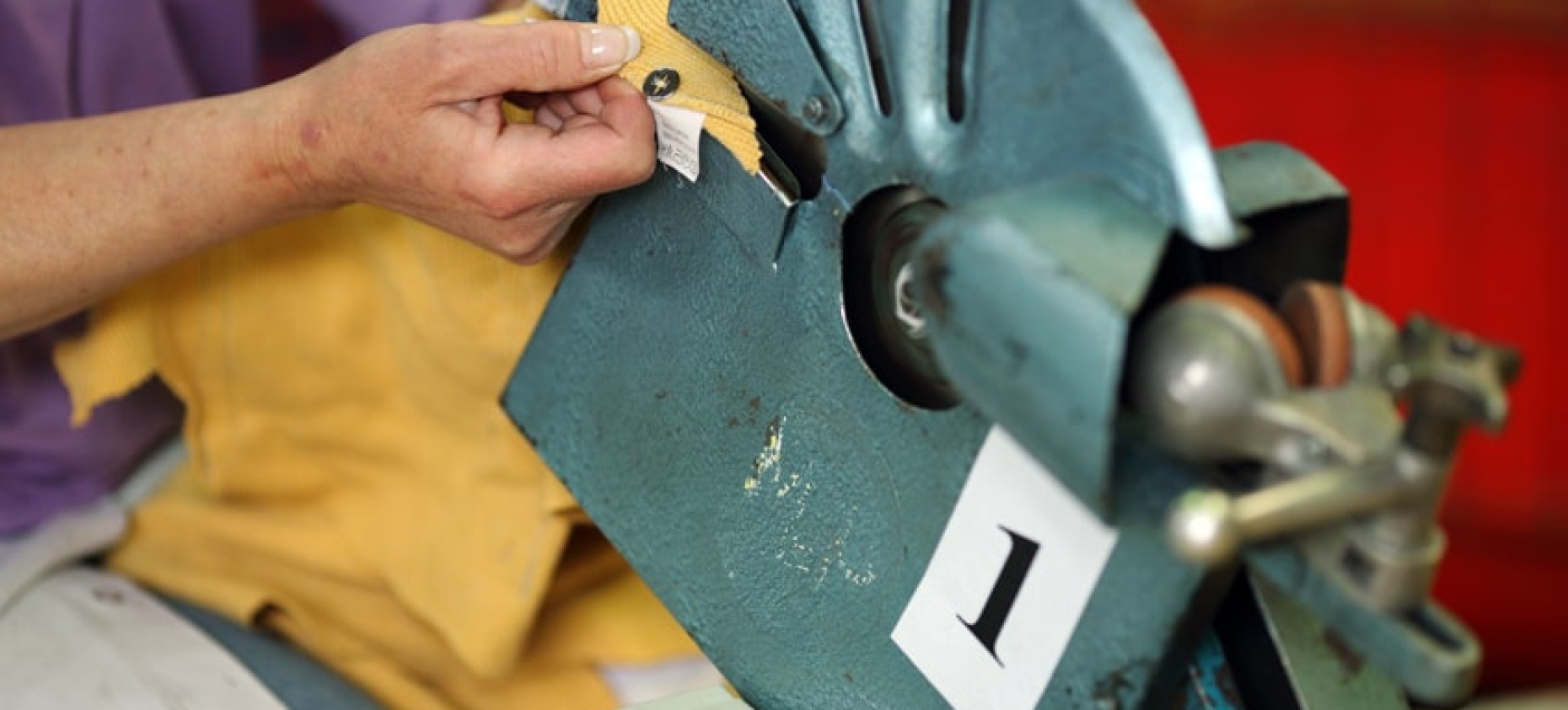Une personne utilise une machine industrielle en métal bleu avec un mécanisme rotatif pour retirer un bouton pression d'un tissu jaune. La machine est étiquetée avec le chiffre "1".
