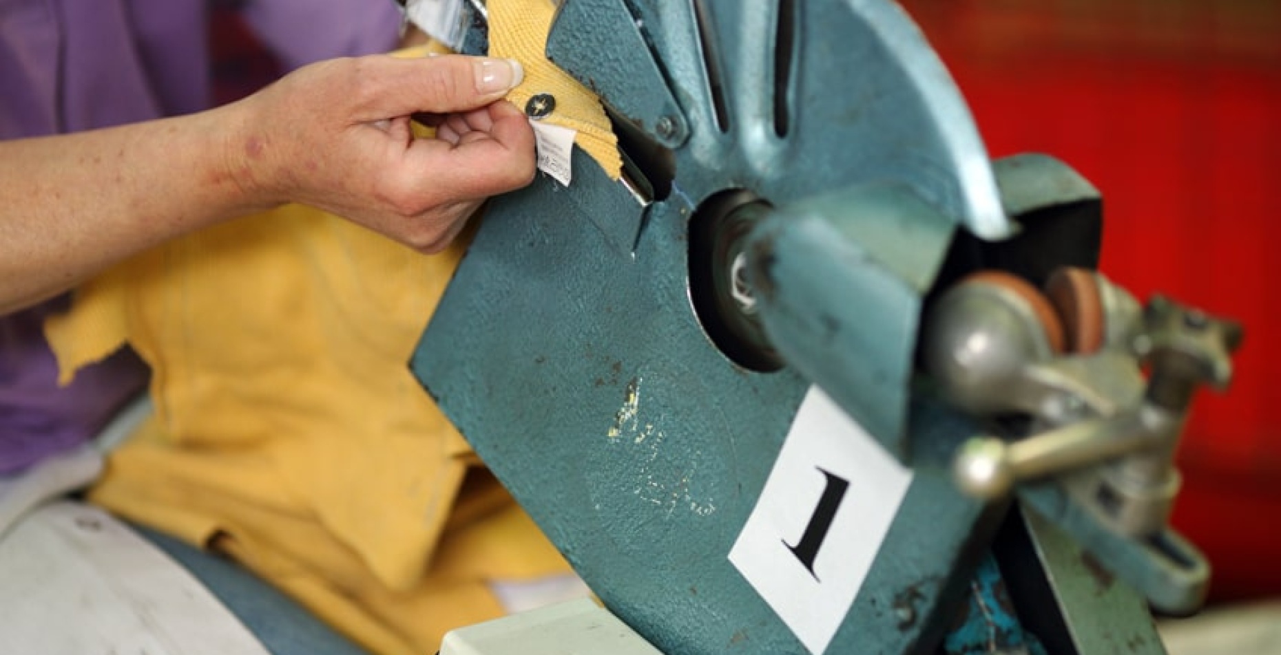 Une personne utilise une machine industrielle en métal bleu avec un mécanisme rotatif pour retirer un bouton pression d'un tissu jaune. La machine est étiquetée avec le chiffre "1".