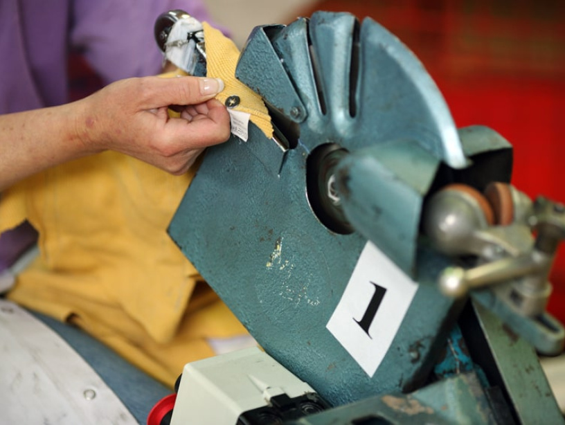 Une personne utilise une machine industrielle en métal bleu avec un mécanisme rotatif pour retirer un bouton pression d'un tissu jaune. La machine est étiquetée avec le chiffre "1".