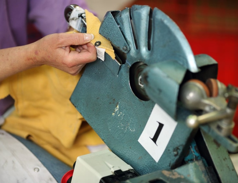 Une personne utilise une machine industrielle en métal bleu avec un mécanisme rotatif pour retirer un bouton pression d'un tissu jaune. La machine est étiquetée avec le chiffre "1".