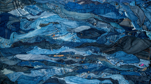 Un amas dense de jeans usagés dans différentes teintes de bleu, formant une texture ondulée et chaotique avec des variations de tissu et des détails comme des poches, coutures et étiquettes visibles.