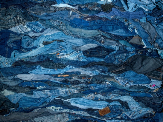 Un amas dense de jeans usagés dans différentes teintes de bleu, formant une texture ondulée et chaotique avec des variations de tissu et des détails comme des poches, coutures et étiquettes visibles.