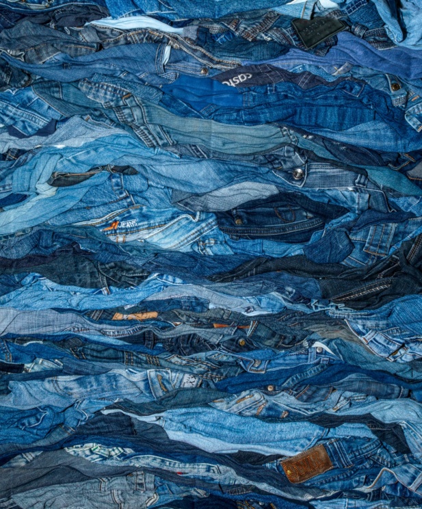 Un amas dense de jeans usagés dans différentes teintes de bleu, formant une texture ondulée et chaotique avec des variations de tissu et des détails comme des poches, coutures et étiquettes visibles.