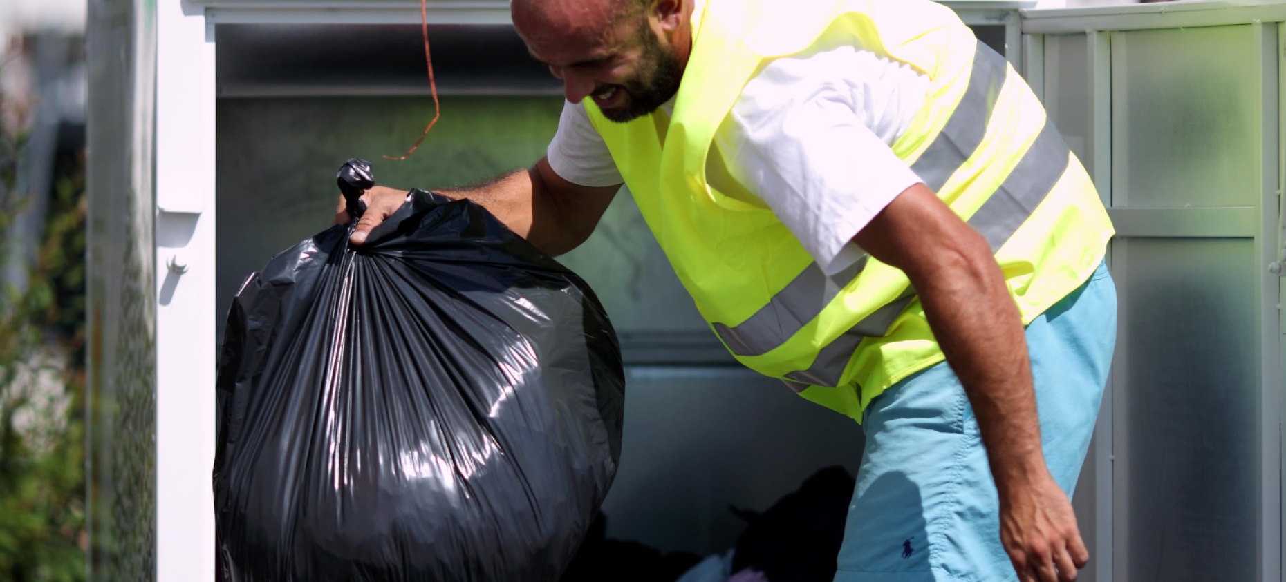Un employé en gilet fluorescent vide un conteneur de collecte de vêtements, triant des sacs remplis d'habits donnés, en extérieur.