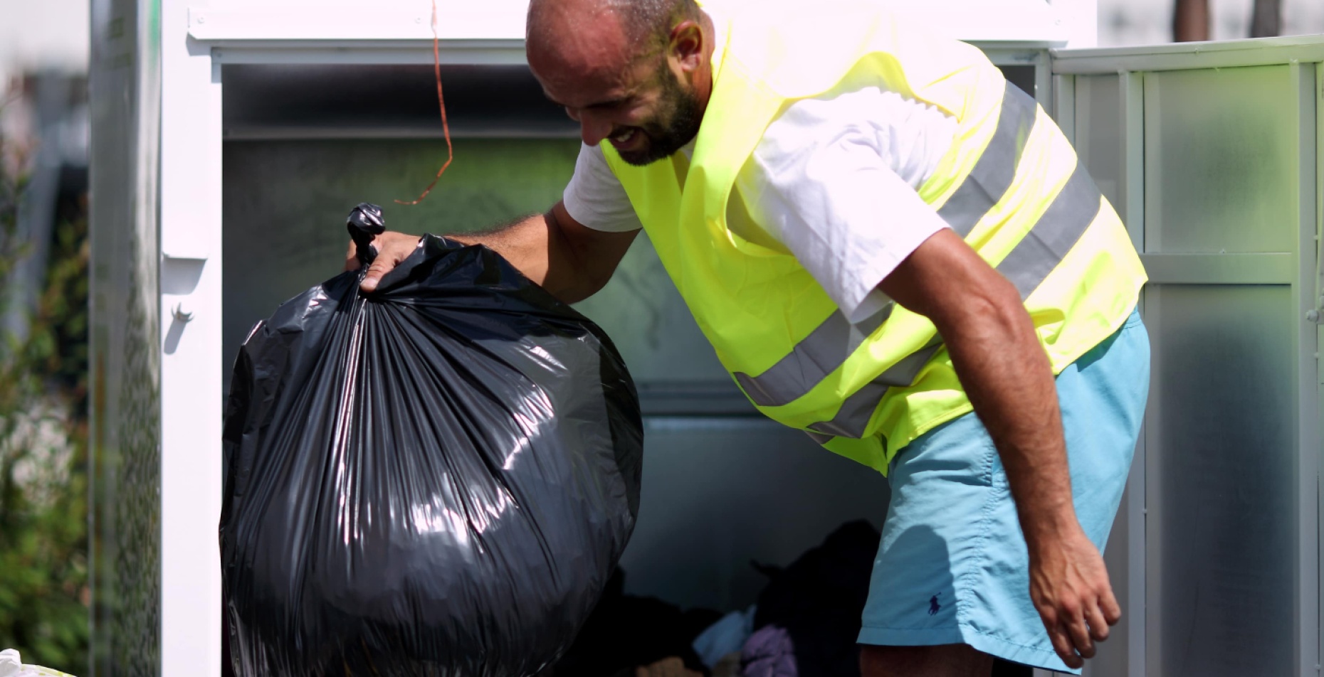 Un employé en gilet fluorescent vide un conteneur de collecte de vêtements, triant des sacs remplis d'habits donnés, en extérieur.