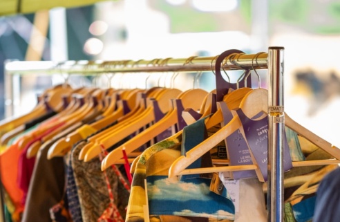 Un portant rempli de vêtements colorés sur des cintres en bois, avec un miroir et une tente en arrière-plan, dans un marché de seconde main.