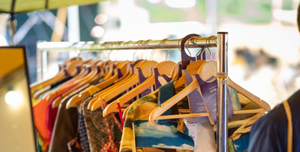 Un portant rempli de vêtements colorés sur des cintres en bois, avec un miroir et une tente en arrière-plan, dans un marché de seconde main.