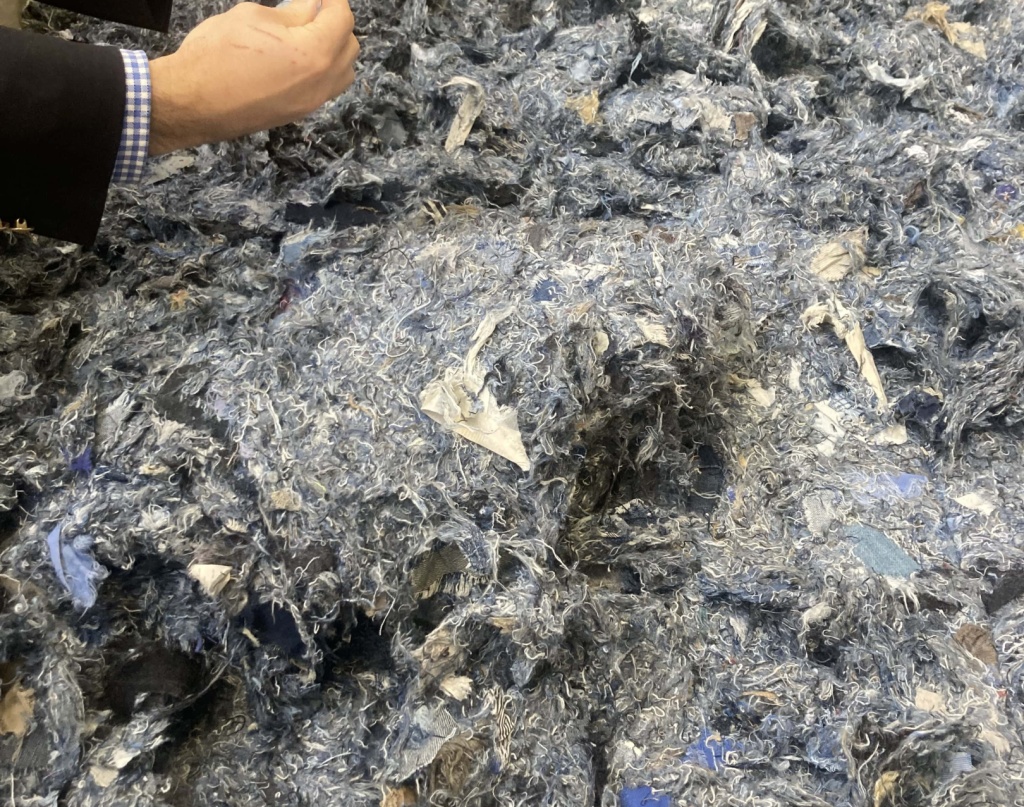 Un amas de fibres textiles recyclées aux teintes majoritairement bleues et grises, composé de résidus de jeans déchiquetés, manipulé par une personne en costume.