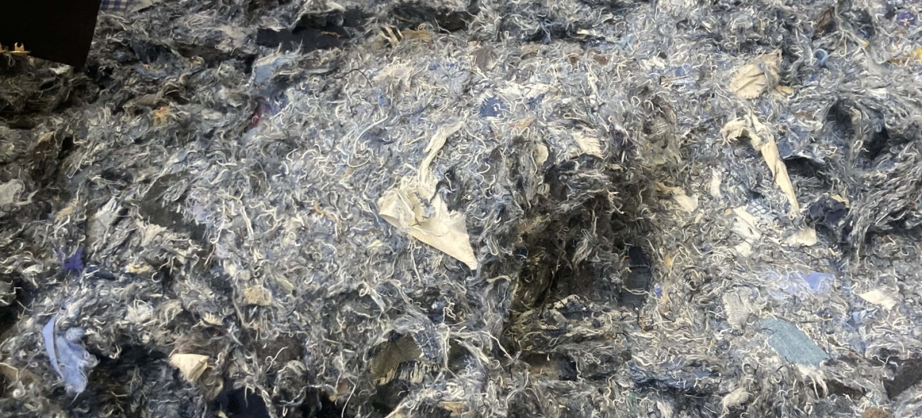 Un amas de fibres textiles recyclées aux teintes majoritairement bleues et grises, composé de résidus de jeans déchiquetés, manipulé par une personne en costume.