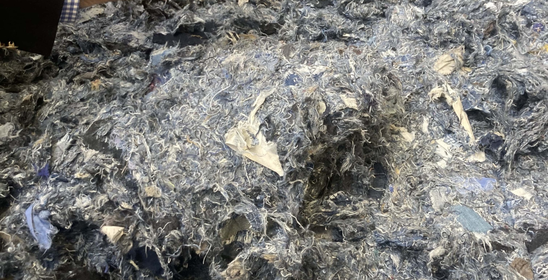 Un amas de fibres textiles recyclées aux teintes majoritairement bleues et grises, composé de résidus de jeans déchiquetés, manipulé par une personne en costume.