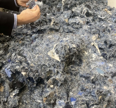 Un amas de fibres textiles recyclées aux teintes majoritairement bleues et grises, composé de résidus de jeans déchiquetés, manipulé par une personne en costume.