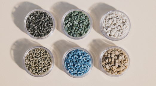 Six récipients transparents contenant des granulés de plastique recyclé aux teintes variées – noir, vert, blanc, beige, bleu et gris – soigneusement disposés sur une surface neutre.