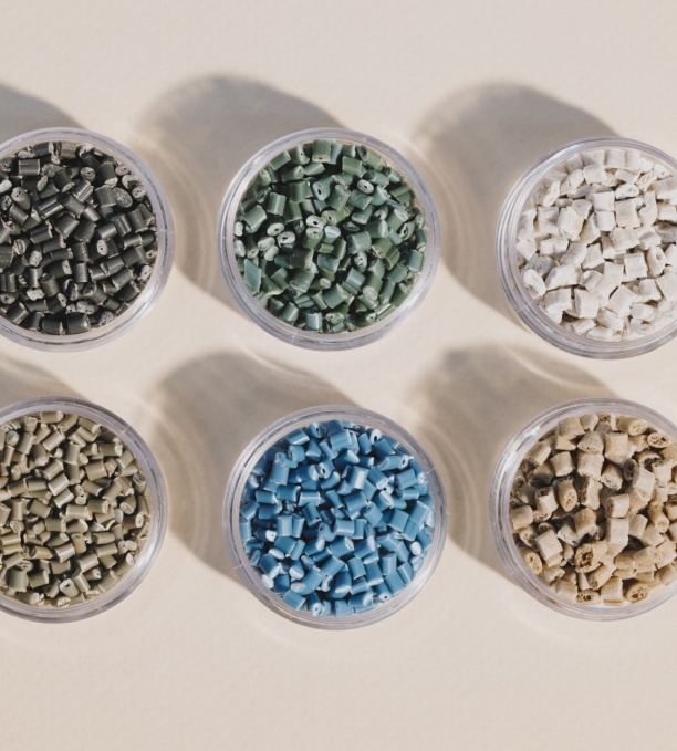 Six récipients transparents contenant des granulés de plastique recyclé aux teintes variées – noir, vert, blanc, beige, bleu et gris – soigneusement disposés sur une surface neutre.
