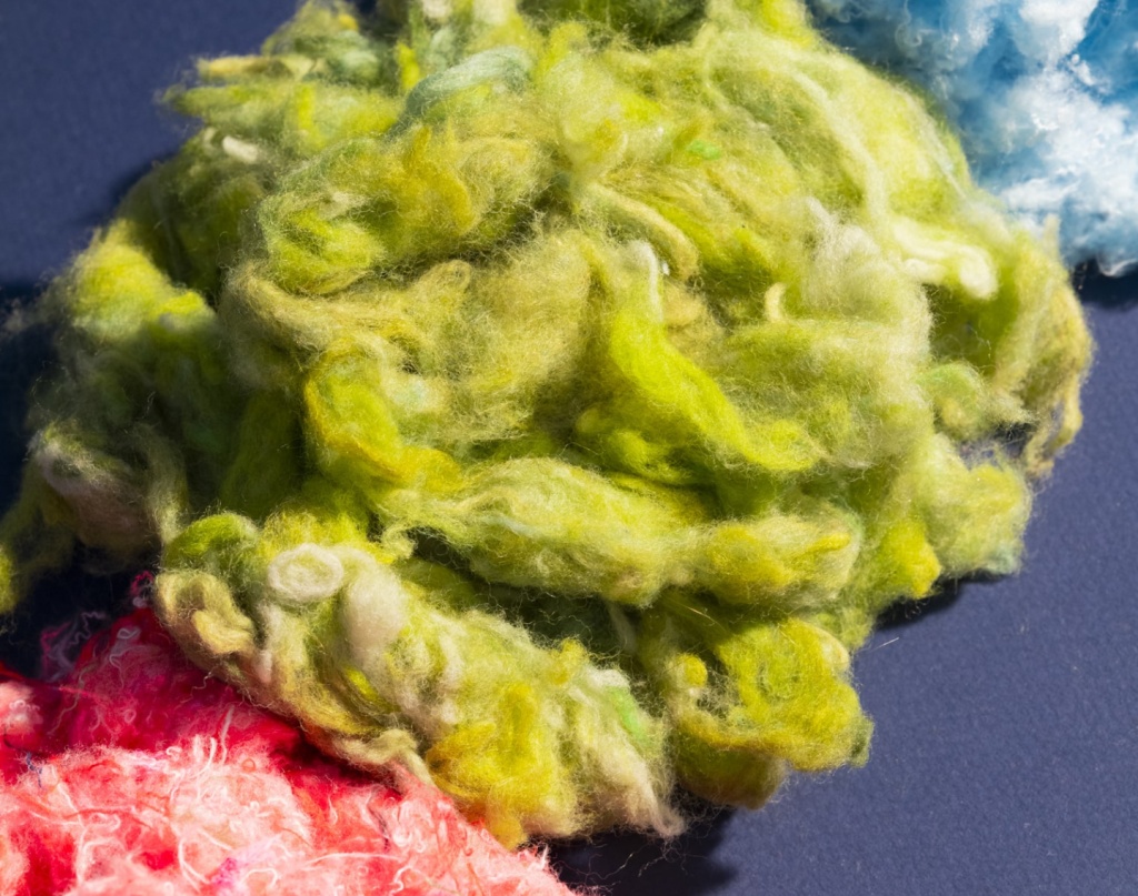 Fibres textiles recyclées aux couleurs vives – vert, bleu et rose – disposées sur un fond sombre, illustrant le processus de transformation des anciens vêtements en nouvelles matières premières.