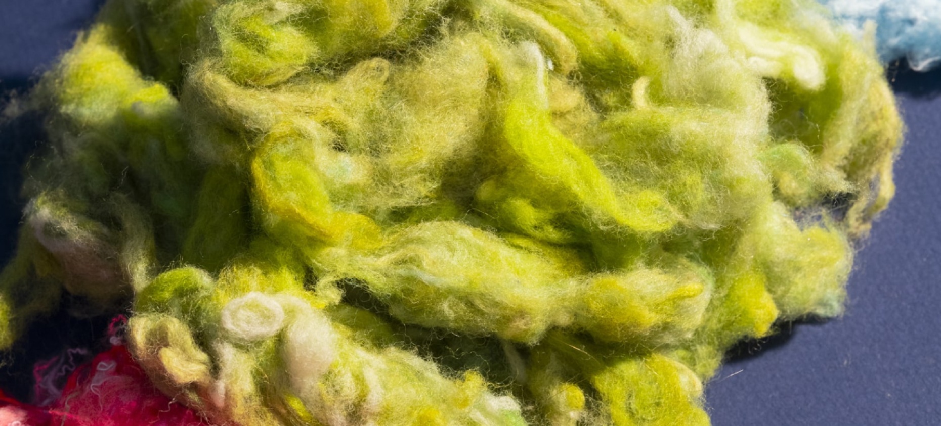 Fibres textiles recyclées aux couleurs vives – vert, bleu et rose – disposées sur un fond sombre, illustrant le processus de transformation des anciens vêtements en nouvelles matières premières.