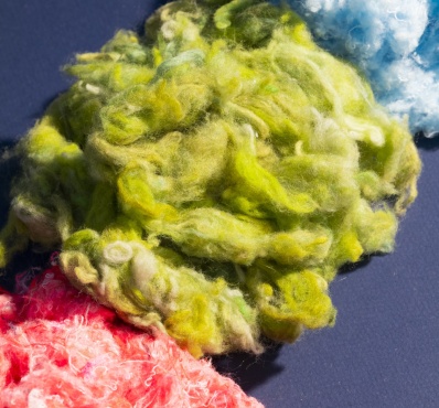Fibres textiles recyclées aux couleurs vives – vert, bleu et rose – disposées sur un fond sombre, illustrant le processus de transformation des anciens vêtements en nouvelles matières premières.