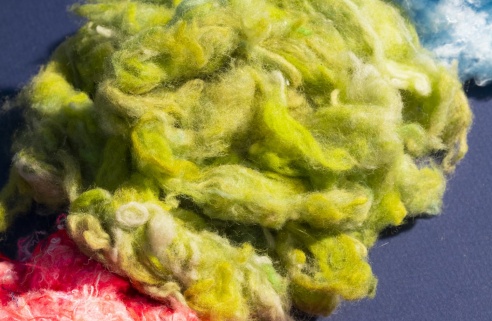 Fibres textiles recyclées aux couleurs vives – vert, bleu et rose – disposées sur un fond sombre, illustrant le processus de transformation des anciens vêtements en nouvelles matières premières.