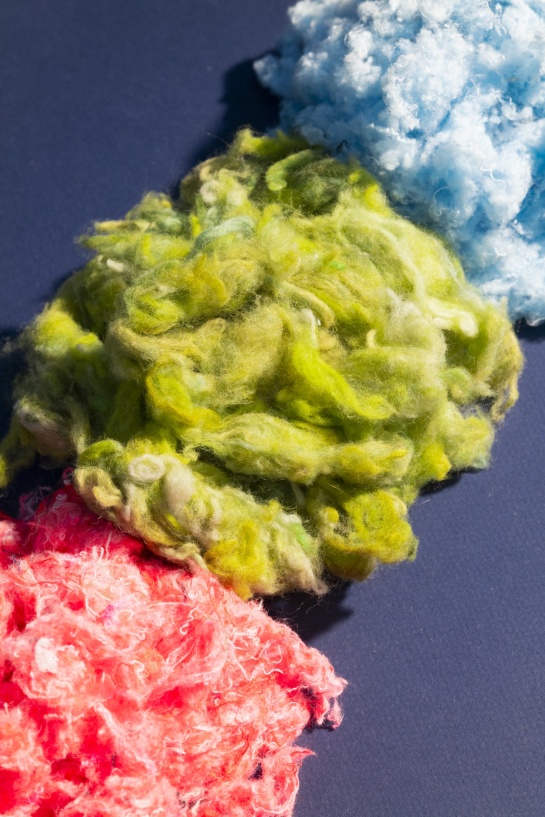 Fibres textiles recyclées aux couleurs vives – vert, bleu et rose – disposées sur un fond sombre, illustrant le processus de transformation des anciens vêtements en nouvelles matières premières.