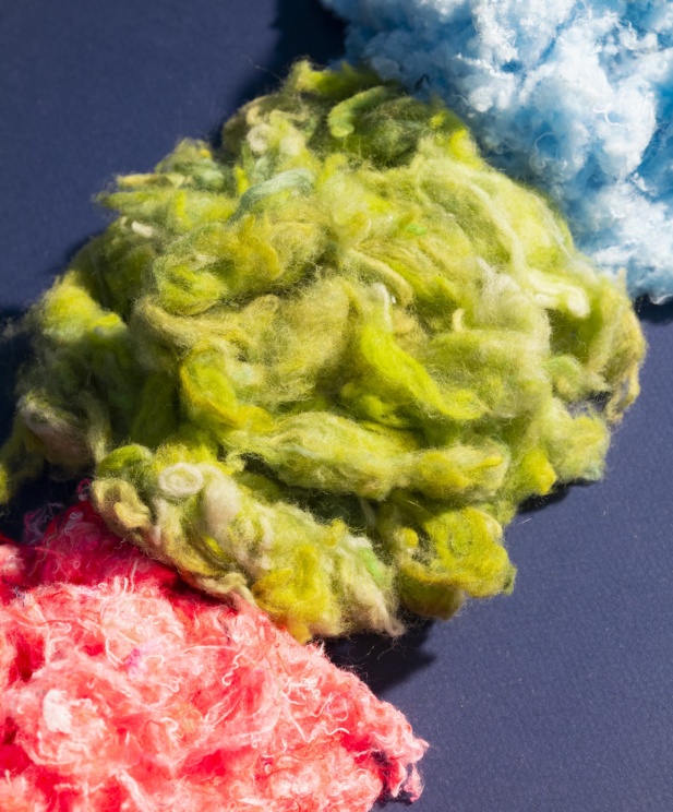 Fibres textiles recyclées aux couleurs vives – vert, bleu et rose – disposées sur un fond sombre, illustrant le processus de transformation des anciens vêtements en nouvelles matières premières.