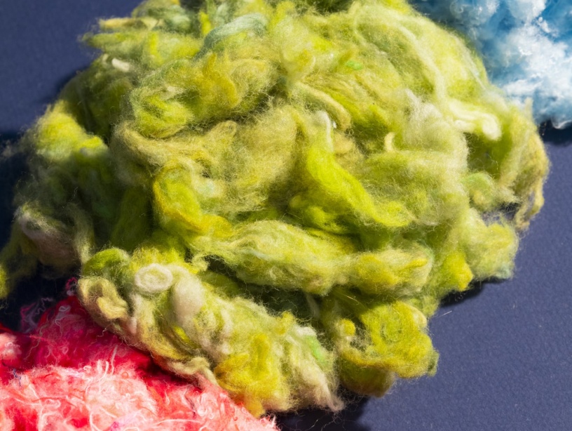 Fibres textiles recyclées aux couleurs vives – vert, bleu et rose – disposées sur un fond sombre, illustrant le processus de transformation des anciens vêtements en nouvelles matières premières.