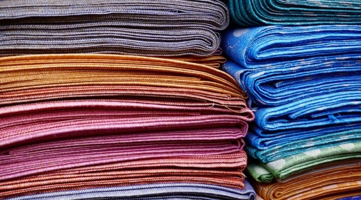 Une pile de tissus soigneusement pliés dans différentes couleurs vibrantes, allant du bleu au doré en passant par le violet et le vert, avec des textures et motifs variés.