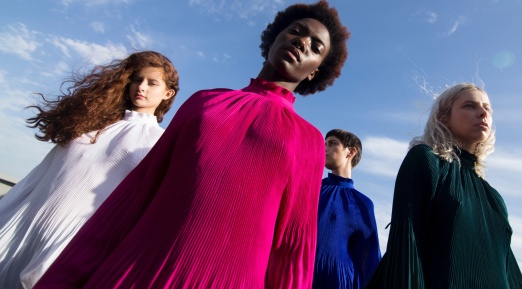 L’image montre quatre femmes portant de longues robes plissées de couleurs vives : fuchsia, blanc, bleu et vert foncé. Elles sont photographiées en contre-plongée sous un ciel bleu avec quelques nuages. Les robes sont fluides et semblent bouger légèrement avec le vent.