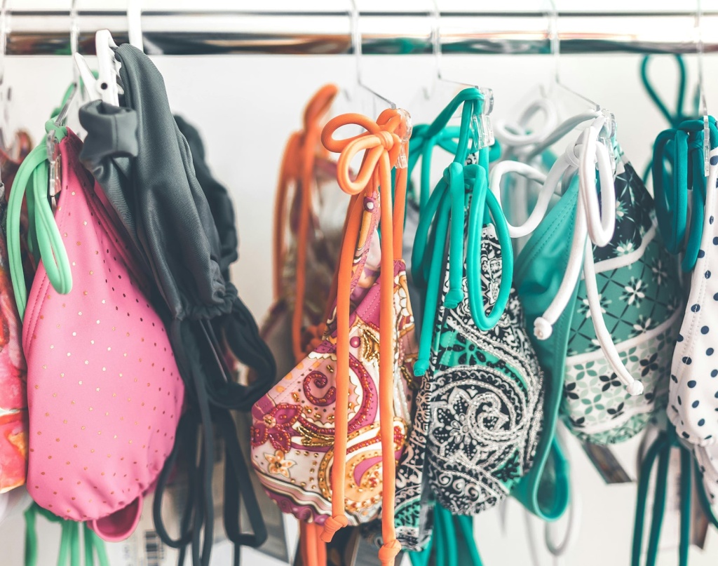 L’image montre un portant rempli de maillots de bain colorés suspendus à des cintres. Les motifs variés et les couleurs vives suggèrent un mélange de styles et de designs, probablement dans un magasin ou un espace de seconde main. L’ensemble dégage une ambiance estivale et dynamique.