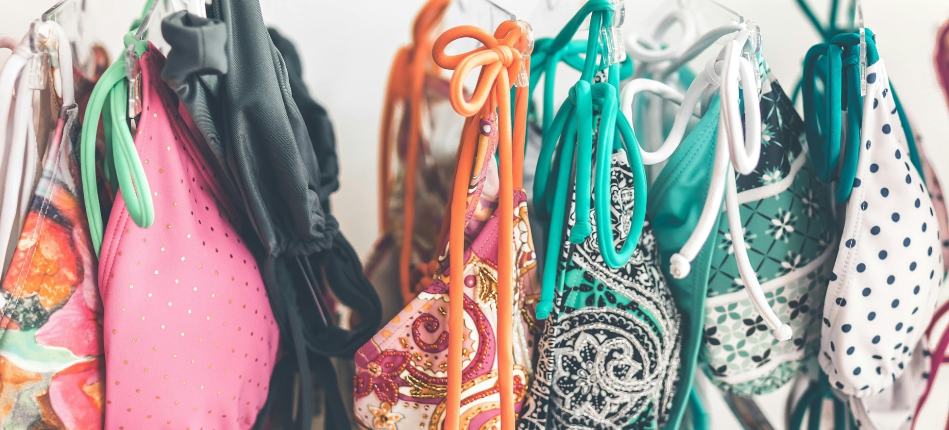 L’image montre un portant rempli de maillots de bain colorés suspendus à des cintres. Les motifs variés et les couleurs vives suggèrent un mélange de styles et de designs, probablement dans un magasin ou un espace de seconde main. L’ensemble dégage une ambiance estivale et dynamique.