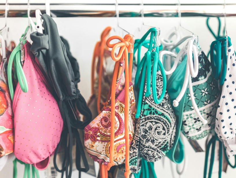 L’image montre un portant rempli de maillots de bain colorés suspendus à des cintres. Les motifs variés et les couleurs vives suggèrent un mélange de styles et de designs, probablement dans un magasin ou un espace de seconde main. L’ensemble dégage une ambiance estivale et dynamique.