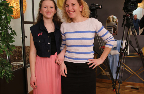 Deux femmes souriantes posent ensemble, complices, devant un décor de studio avec des caméras. L’une porte un haut rayé bleu et blanc, l’autre une veste sans manches avec l’inscription « COOL ».