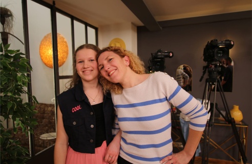 Deux femmes souriantes posent ensemble, complices, devant un décor de studio avec des caméras. L’une porte un haut rayé bleu et blanc, l’autre une veste sans manches avec l’inscription « COOL ».