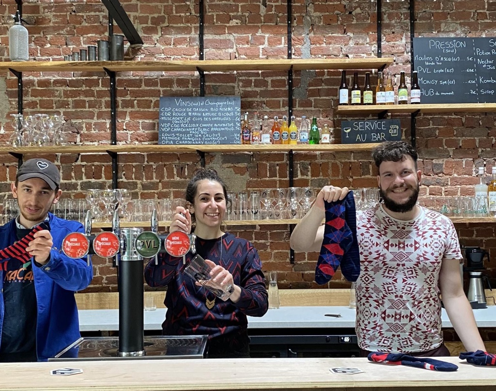 L’image montre trois personnes souriantes derrière un bar avec un fond en briques apparentes. Ils tiennent et présentent des chaussettes colorées avec des motifs. L’ambiance semble chaleureuse et conviviale, évoquant un lieu accueillant où se mêlent artisanat, commerce local et bonne humeur.