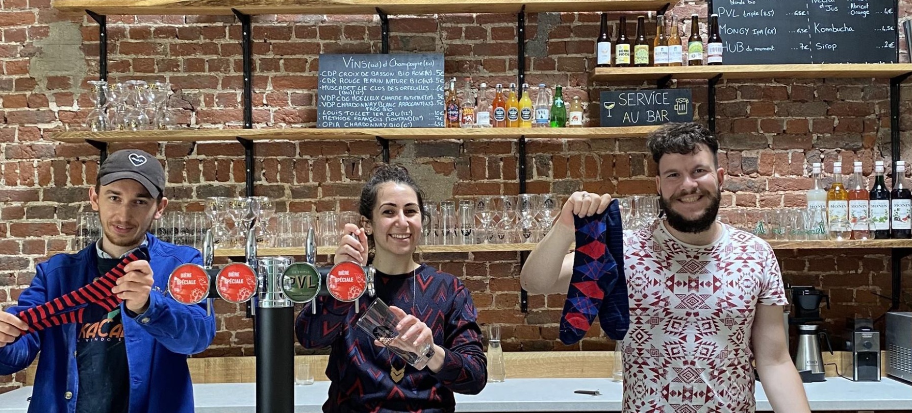 L’image montre trois personnes souriantes derrière un bar avec un fond en briques apparentes. Ils tiennent et présentent des chaussettes colorées avec des motifs. L’ambiance semble chaleureuse et conviviale, évoquant un lieu accueillant où se mêlent artisanat, commerce local et bonne humeur.