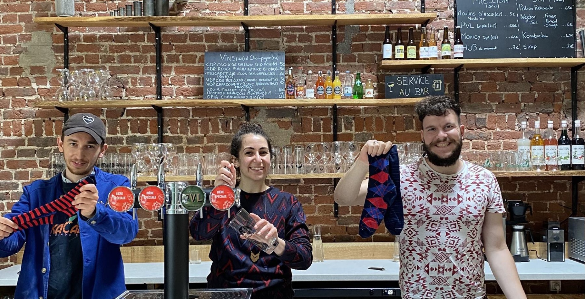 L’image montre trois personnes souriantes derrière un bar avec un fond en briques apparentes. Ils tiennent et présentent des chaussettes colorées avec des motifs. L’ambiance semble chaleureuse et conviviale, évoquant un lieu accueillant où se mêlent artisanat, commerce local et bonne humeur.